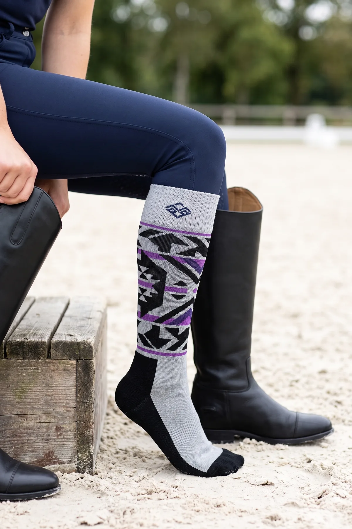 Chaussette d’équitation haute à motif jacquard et logo dans le revers, portée sur un pantalon marine à côté d’une botte